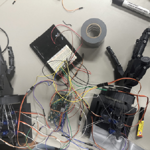 A complex breadboard configuration is shown on a table, connected to two robotic hands via numerous colored jumper wires. The setup includes a microcontroller and multiple servo motors, with duct tape and tools scattered around